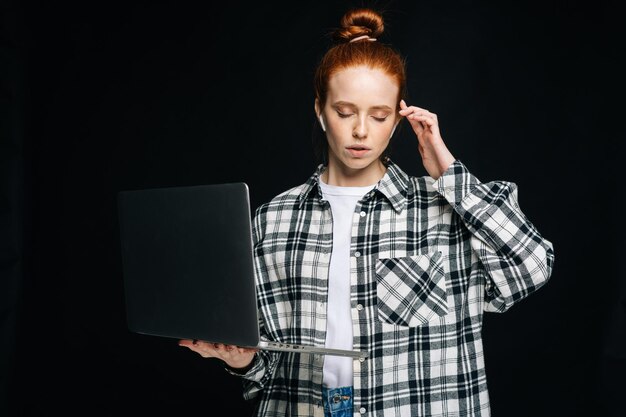 Overworked sad young woman student having severe headache while working by laptop computer on isolated black background Pretty redhead lady model emotionally showing facial expressions in studio