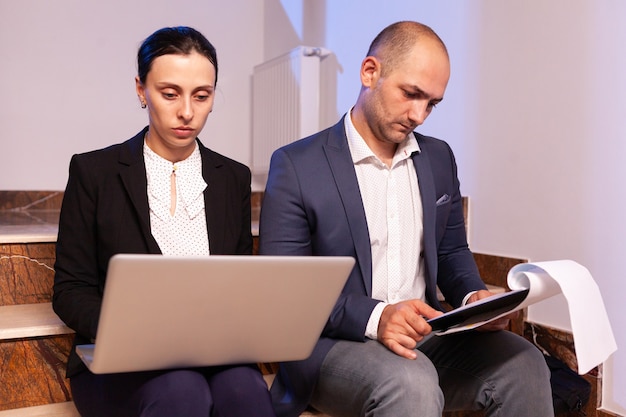 Overworked tired businessman doing overtime sitting on stairs helping coworker with diffcult deadline. Colleague entrepreneur working late together in evening at corporate job sitting on staircase, ex