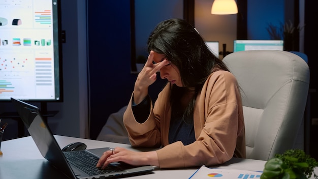 Donna d'affari stanca oberata di lavoro che sfrega la faccia lavorando alle statistiche di gestione sul computer portatile a tarda notte nell'ufficio di avvio. imprenditore esausto che controlla la scadenza della società di grafica economica