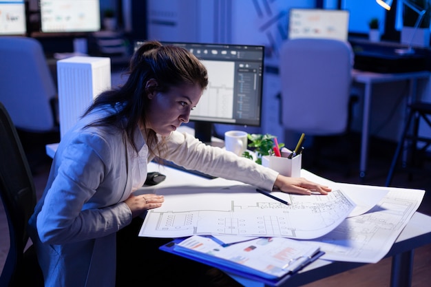 Overworked woman architect checking and matching blueprints sitting at office desk in front of computer