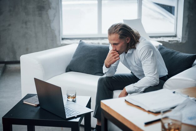 Overworked young businessman sitting on couch and watching at laptop