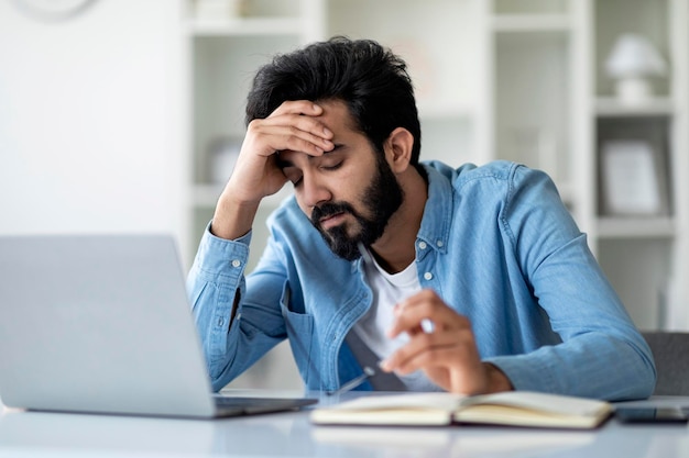 Overworking tired indian man sitting at desk with laptop and touching head