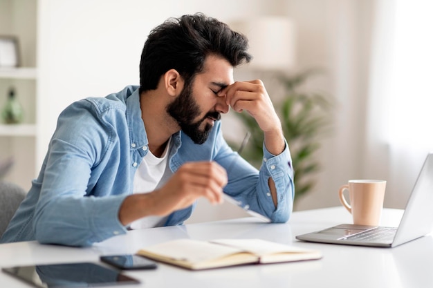 Overworking tired indian man suffering eyes strain while using laptop at home