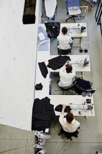 Overzicht van drie jonge vrouwen die door elektrische naaimachines in werkplaats zitten tijdens hun werk over seizoensmodecollectie