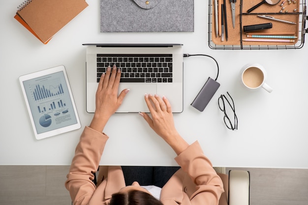 Overzicht van een jonge elegante zakenvrouw die bij een bureau voor een laptop zit en toetsen aanraakt tijdens het invoeren van nieuwe gegevens of netwerken