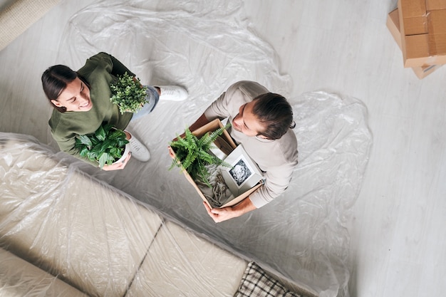 Overzicht van gelukkige jonge man en vrouw met groene binnenlandse planten die op de vloer staan door de bank in de woonkamer