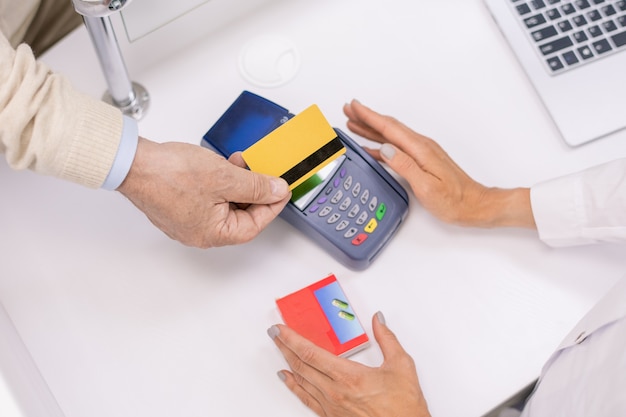 Overzicht van handen van senior koper die creditcard over het scherm van de betaalterminal houdt terwijl hij voor medicijnen betaalt aan de kassa