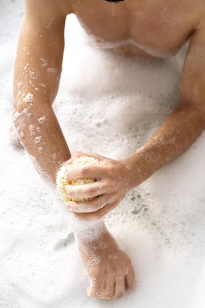 Foto overzicht van shirtless jongeman met spons zijn armen wassen zittend in bad met warm water en schuim