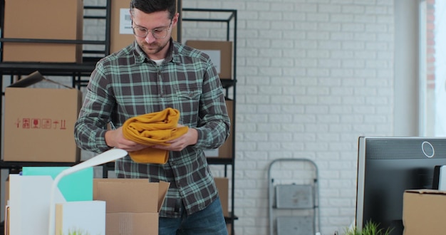 The owner of an online store works on a computer at a desk in a warehouse A male worker is packing a yellow jumper in a room with shelves full of parcels