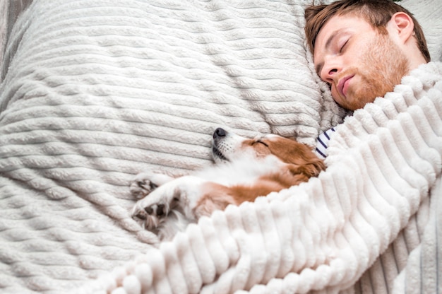 飼い主は犬を毛布の下でベッドに寝かせて寝ます。白色の背景。コンセプトウィークエンド。睡眠と休息の概念。コピースペース