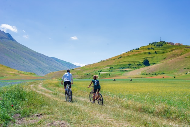 Paar fietsen mtb in bloeiende gecultiveerde velden, beroemde kleurrijke bloeiende vlakte in de Apennijnen, Castelluccio di Norcia hooglanden, Italië. Linzen, rode klaprozen en blauwe korenbloemen.