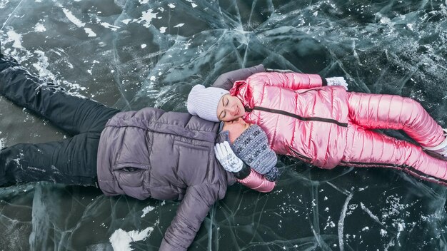 Paar heeft plezier winterwandeling tegen de achtergrond van ijs van bevroren meer Liefhebbers liggen op helder ijs met scheuren veel plezier kus en knuffel Uitzicht van bovenaf Gelukkige mensen op besneeuwd ijs Huwelijksreis liefdesverhaal