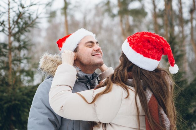 Paar in een kerstmuts en knuffelen en gek doen in een winter naaldbos