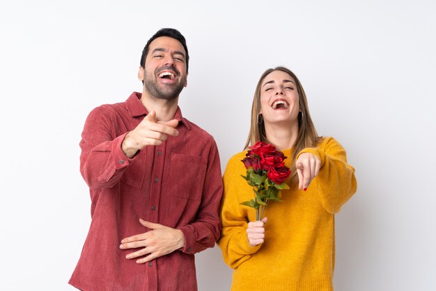 Paar in Valentine Day-holdingsbloemen over geïsoleerde muur die met vinger op iemand richten en veel lachen