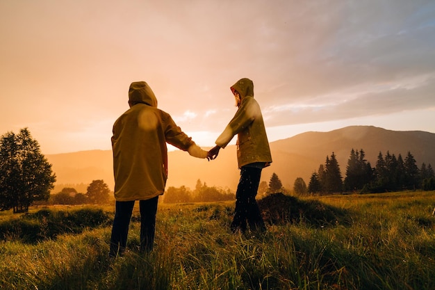 Paar jonge wandelaars in gele regenjassen die op een weide in de bergen staan, hand in hand