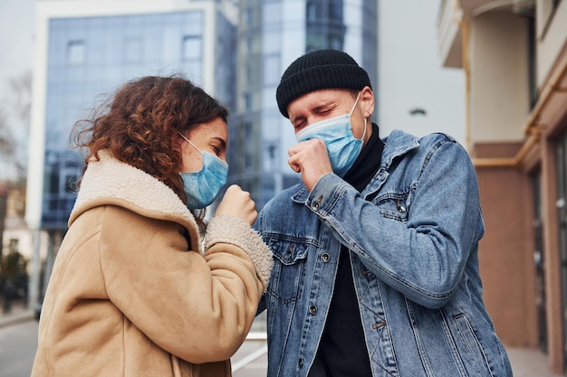 Paar met beschermende maskers hoesten buiten in de stad in de buurt van bedrijfsgebouw in quarantainetijd Conceptie van coronavirus