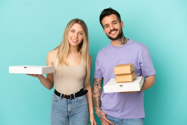 Paar met pizza's en hamburgers over geïsoleerde blauwe achtergrond poseren met armen op heup en glimlachend