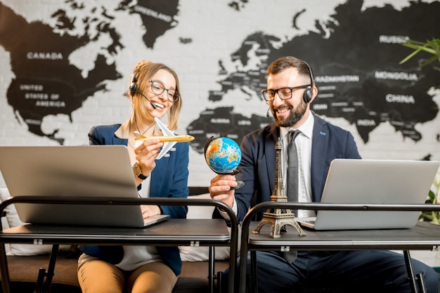 Foto paar reismanagers spelen met wereldbol en vliegtuig zittend op het bureau van het agentschap met wereldkaart op de achtergrond