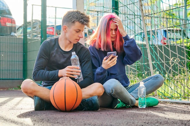 Paar tieners op straat sportveld met bal, levensstijl, jongen en meisje 16 jaar oud praten, drinkwater uit fles kijken naar smartphone scherm