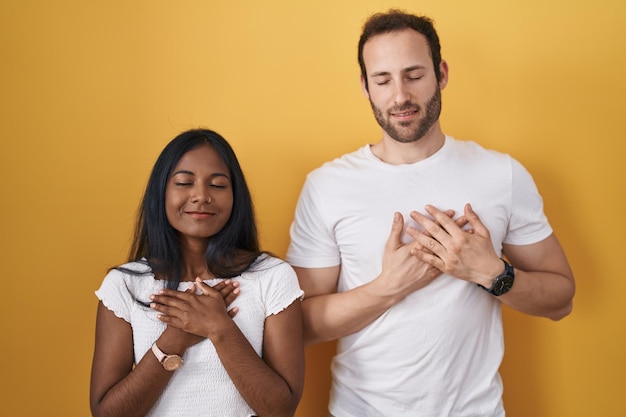 Paar tussen verschillende rassen dat zich over gele achtergrond bevindt glimlachend met handen op borst met gesloten ogen en dankbaar gebaar op het concept van de gezichtsgezondheid