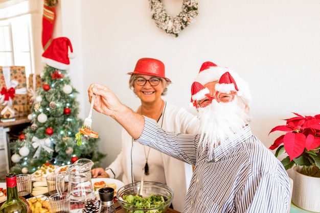 Paar twee gelukkige senioren die de kerstdag eten en plezier hebben tijdens de lunch - kerstboom op de achtergrond - glimlachen en lachen samen aan tafel - oude man met een baardmasker genieten