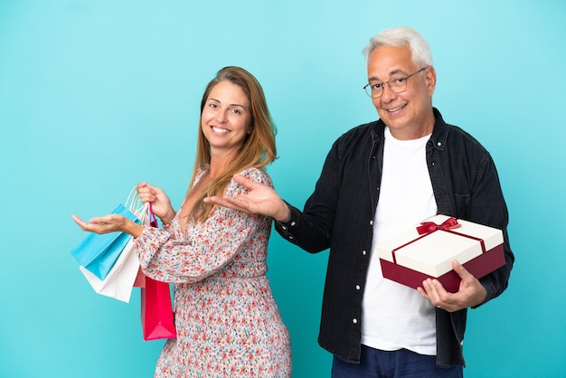 Paar van middelbare leeftijd met boodschappentas en cadeau geïsoleerd op een blauwe achtergrond die de handen naar de zijkant uitstrekt om uit te nodigen om te komen