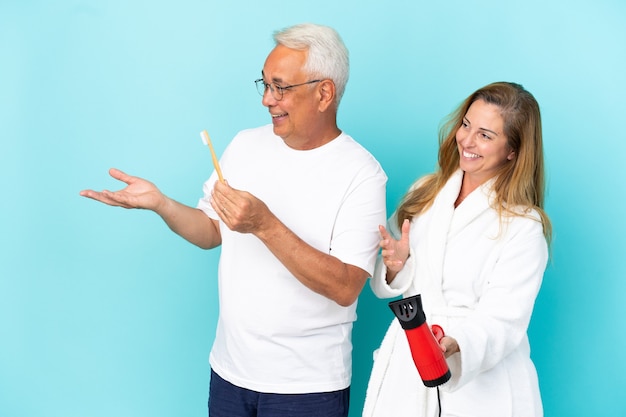 Paar van middelbare leeftijd met droger en tandenborstel geïsoleerd op een blauwe achtergrond die naar achteren wijst en een product presenteert