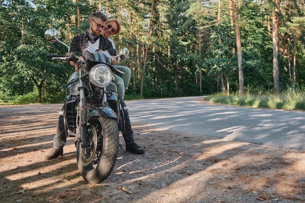 Paar van middelbare leeftijd praten en plezier hebben zittend op een motorfiets die samen op een bosweg reist