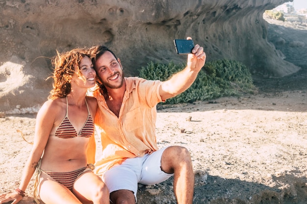 Paar volwassenen op het strand praten en kijken naar de telefoon van de vrouw die op de rotsen zit - vrouw in bikini die naar hun telefoon kijkt en een man die naar dezelfde telefoon kijkt