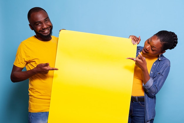 Paar wijzend op blanco kartonnen papier over blauwe achtergrond. Mensen die verliefd zijn, houden een mockup-uithangbord vast om een sms-bericht voor advertenties in de studio te plaatsen. Man en vrouw met vrije ruimtebanner.