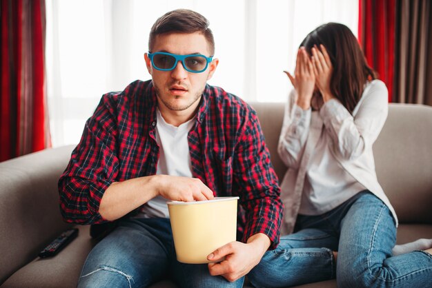 Paar zittend op de bank, man in 3d-bril met popcorn in handen film kijken, bange vrouw sluit haar gezicht met handen