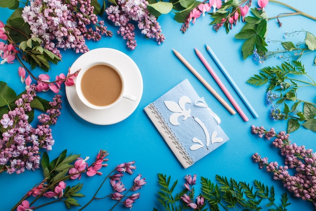 Paarse lila en bloeden hart bloemen en een kopje koffie met laptop en kleurpotloden op pastel blauwe achtergrond