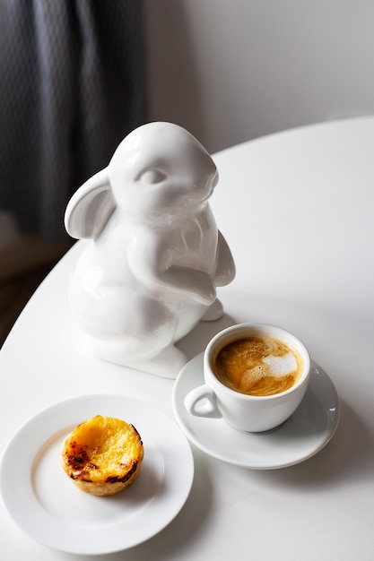 Paashaas staat in de buurt van een kopje cappuccino en een cake op een bord een witte kop koffie Paastafeldecoratie kopieerruimte