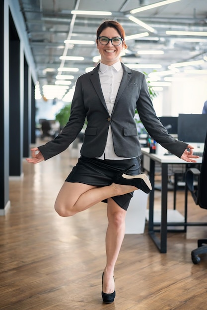 A pacified business woman doing yoga in an open space office A redhaired smiling female employee in a casual suit and glasses stands in a tree pose at the workplace Meditation and relaxation