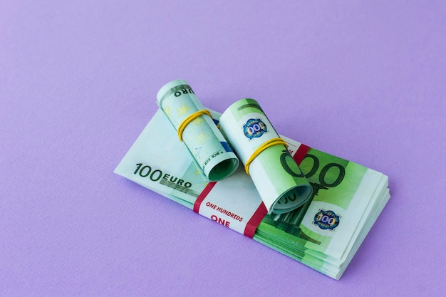 A pack of 100 euro banknotes in a bank package and two rolls\
under an elastic band on it. lilac background.