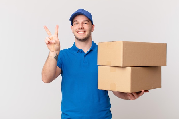 Package deliver man smiling and looking happy, carefree and positive, gesturing victory or peace with one hand