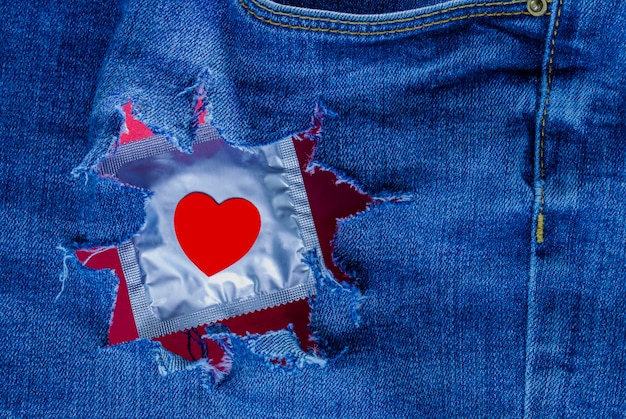 Photo packaged condom with a red heart in torn denim pants. love and romance. safe sex. sex offer. valentine's day card.