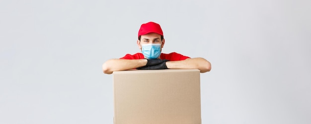 Photo packages and parcels delivery covid19 quarantine and transfer orders young courier in red uniform cap face mask and gloves leaning on box to deliver shipping your orders grey background