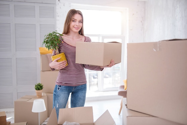 packing actively. beautiful young woman standing in the middle of the living room and smiling at the camera while carrying a carton box and a plant