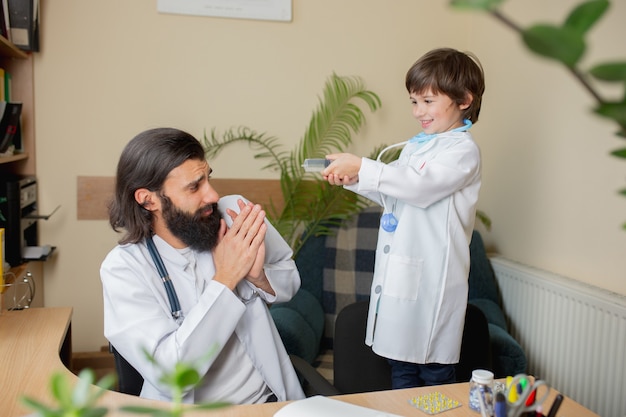 Foto medico pediatra che esamina un bambino in uno studio medico confortevole. concetto di assistenza sanitaria, infanzia, medicina, protezione e prevenzione. il ragazzino si fida del dottore. divertirsi mentre si prende la medicina