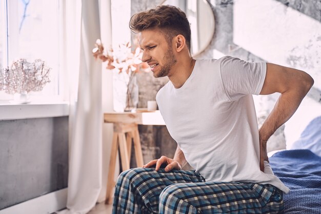 Pain in back. Upset bearded male person touching his back while sitting on the bed