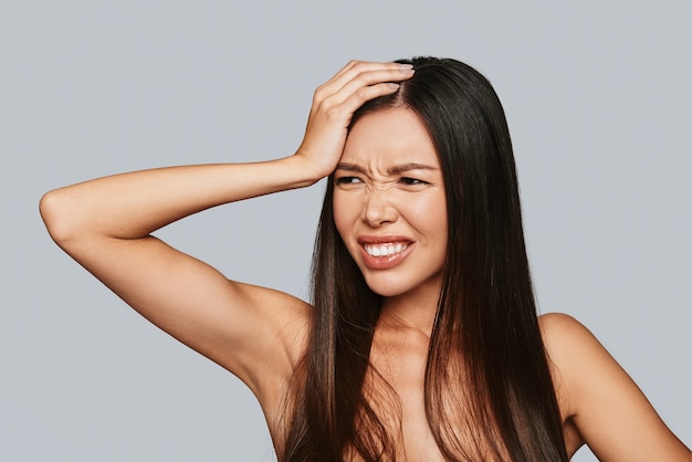 In pain. Frustrated young Asian woman making a face and keeping hand on head while standing against grey background