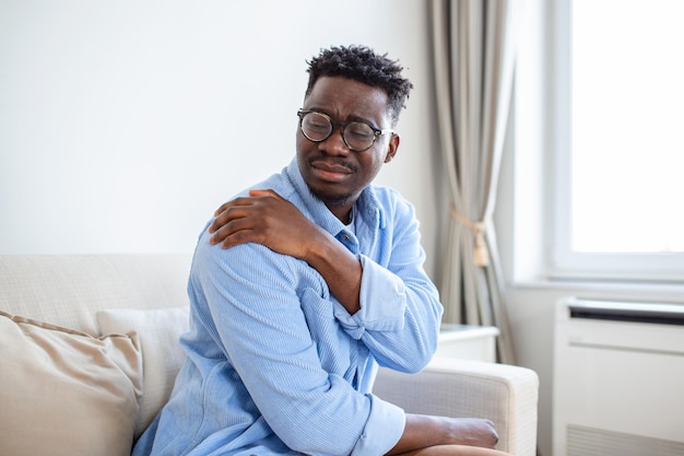 Pain in his shoulder Upper arm pain People with bodymuscles problem Healthcare And Medicine concept Attractive young man sitting on the sofa and holding painful shoulder with another hand