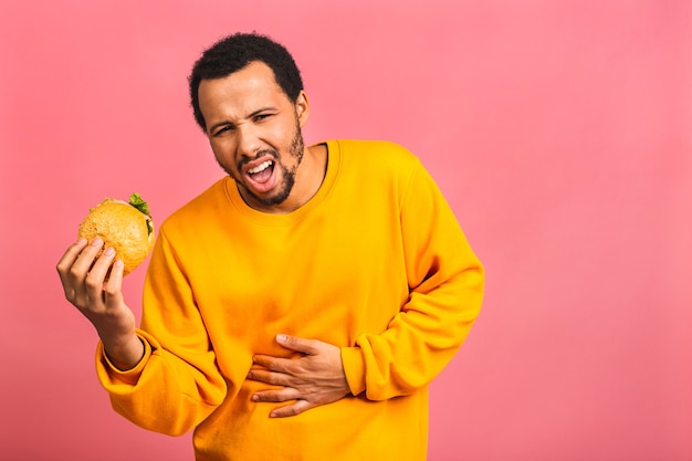 Pain in stomach, abdominal pain. Young man eating hamburger isolated over pink