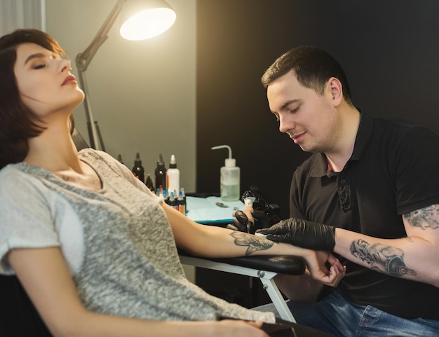 Painful tattooing process, young woman enduring pain while master making tattoo on her arm. Popular body modification, modern lifestyle, copy space