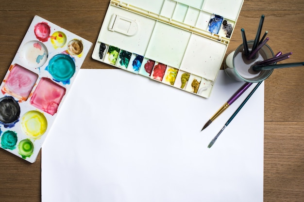 Paint brush on white paper with colorful watercolor and equipment on wooden background.