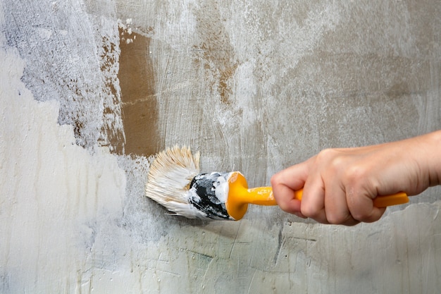 Кисть с желтой пластиковой ручкой в руке художника, перекрашивающего зеленую стену в белый цвет.