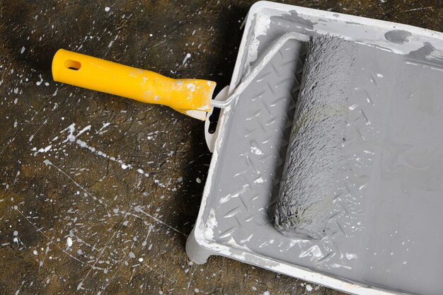 Paint roller with yellow handle in tray for roller with gray\
paint on concrete floor