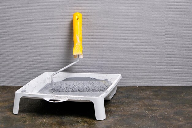 Paint roller with yellow handle in tray for roller with gray\
paint on grey painted wall background