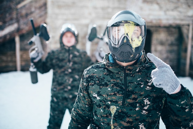 Paintballspeler wijst vinger naar spetterde masker, team na winterse strijd. Extreme sportgame, soldaten in speciaal uniform, paintball